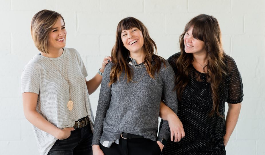 3 women smiling and standing beside white wall